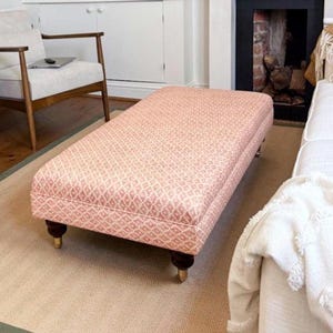 May include: A rectangular ottoman upholstered in a pink and white geometric fabric. The ottoman sits on dark brown legs with gold-toned feet. It is placed on a beige rug with a green border, near a white sofa and a wooden chair.