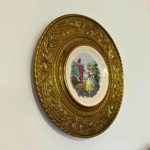 May include: A round decorative wall plate featuring a central ceramic insert with a colourful illustration of two figures in a garden setting, surrounded by a wide, ornate, embossed brass frame with detailed floral and leaf patterns.