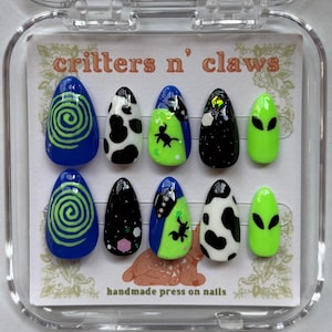 May include: A set of handmade press-on nails in a clear plastic case. The nails feature various designs, including blue swirls, black and white cow print, alien graphics, and a starry night design. The text "critters n' claws" is visible.