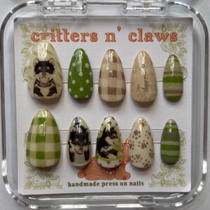 May include: A set of ten press-on nails with various designs. The designs include a dog portrait, green polka dots, brown plaid, and green and brown stripes. The text "critters n' claws" and "handmade press on nails" are visible.