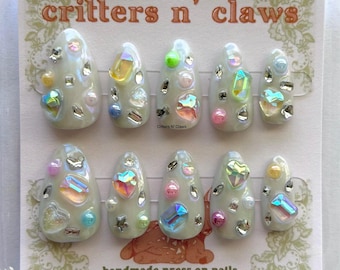 May include: A set of ten almond-shaped press-on nails in a glossy off-white finish, decorated with iridescent rhinestones, colorful beads, and heart-shaped gems. The packaging features the text critters n' claws handmade press on nails.