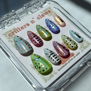 May include: A set of press-on nails in a clear plastic case. The nails are oval-shaped and feature a holographic fish design with black dot and stripe details. The text "critters n' claws" and "handmade press on nails" are visible.