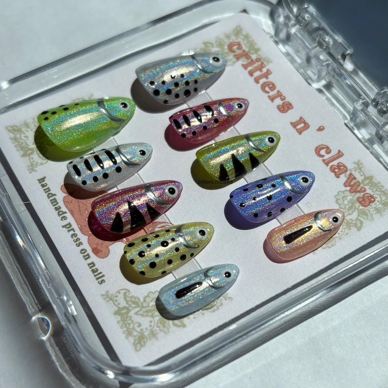May include: A set of handmade press on nails in a clear case. Each nail is shaped like a fish with iridescent colors, including green, pink, silver, and blue. The nails have black dot and stripe details. The text "critters n' claws" is visible.