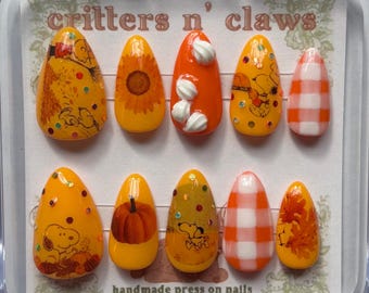 May include: A set of press-on nails with various designs, including Snoopy, sunflowers, pumpkins, and gingham patterns. The nails are primarily orange and yellow, with accents of white and brown. The text "critters n' claws" and "handmade press on nails" are visible.