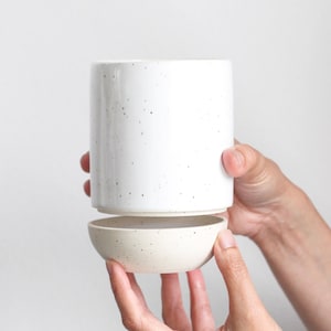 May include: A white ceramic planter with a speckled finish. The planter is divided into two sections, a taller cylinder and a smaller bowl. The planter is being held by two hands.