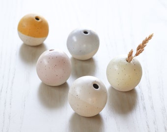 May include: Five small ceramic vases in various colors, including yellow, pink, white, and gray. The vases are round and have a small opening at the top. One vase has a dried stalk of wheat in it.