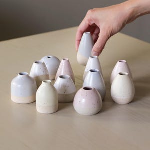 May include: A collection of ten small ceramic vases in various shades of pink, white, and gray. The vases are speckled with small white dots and have a rounded, bulbous shape.