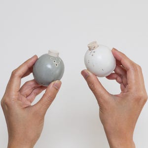 May include: Two ceramic salt and pepper shakers in the shape of spheres. One shaker is gray with a cork top and the other is white with a cork top.