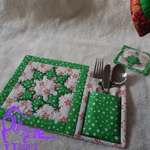 May include: A green and white quilted placemat with a snowflake design and a matching green and white cutlery holder. The placemat has a pocket for holding a fork, knife, and spoon.
