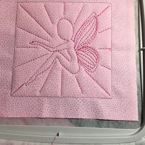 May include: Pink fabric with a white heart pattern and a pink embroidered fairy with wings. The fairy is sitting with its legs crossed and has a sunburst design behind it.