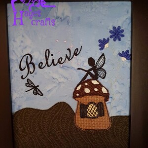 May include: Embroidered artwork featuring a fairy sitting on a mushroom house with the word "Believe" stitched in black thread. The background is a light blue with white stars and a dragonfly.