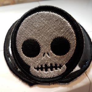 Könnte beinhalten: Ein gestickter Totenkopf-Patch in Schwarz und Weiß mit einem gestickten Lächeln. Der Patch ist von einem schwarzen Rand umgeben.