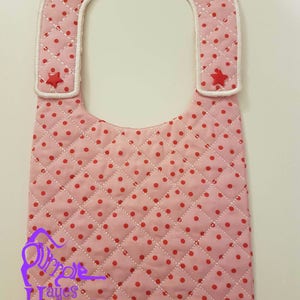May include: Pink quilted baby bib with white trim and red polka dots. The bib has two red star-shaped buttons on the collar.