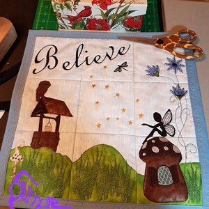 May include: A handmade quilted wall hanging with a blue border and a white background. The centre of the quilt features the word "Believe" in black cursive lettering. The quilt also features a fairy sitting on a mushroom, a well, and a dragonfly. The quilt is signed "Hayes Crafts" in purple lettering.