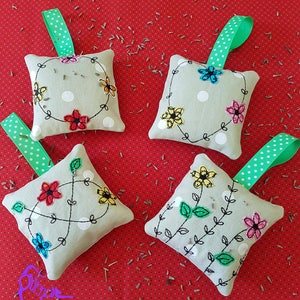 May include: Four handmade fabric sachets with embroidered floral designs. Each sachet has a green and white polka dot ribbon attached. The sachets are on a red background with scattered lavender seeds.