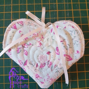 Può includere: Un ornamento di tessuto a forma di cuore bianco con un motivo floreale rosa e un nastro di raso bianco. L'ornamento è trapuntato con un motivo a spirale e ha un fiocco di raso rosa.