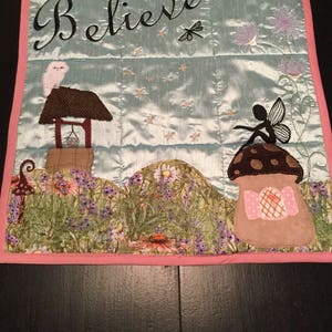 May include: A light blue quilted wall hanging with a pink border. The centre of the hanging features the word "Believe" in black cursive lettering. The hanging also features a whimsical fairy sitting on a mushroom, a small owl perched on a well, and a dragonfly.