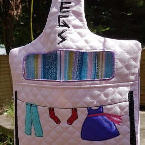 May include: Pink fabric hanging organizer with a quilted design. It has a pocket at the top and a line of clothes hanging on the bottom. The clothes include a pair of blue pants, two red socks, and a purple dress with a pink ribbon.