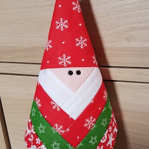 Op de afbeelding: Een rode en groene kerst-gnome-versiering met een witte baard en sneeuwvlokpatronen. De gnome hangt aan een zwart-wit geblokt lint.
