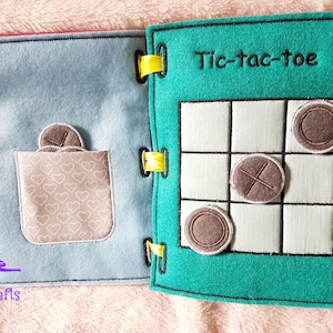 May include: A blue and green felt book with a tic-tac-toe game on one side and a pocket with felt circles on the other side. The text "Tic-tac-toe" is embroidered on the green side.