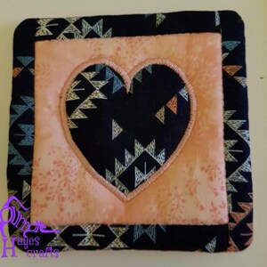 May include: A pink and black quilted coaster with a heart-shaped design. The heart is embroidered with a geometric pattern in shades of blue, white, and orange.
