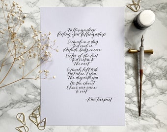 May include: A white sheet of paper featuring black calligraphy of a poem by Kae Tempest, accompanied by a grey and gold calligraphy pen, a small ink pot, gold paper clips, and a sprig of dried white baby's breath flowers.