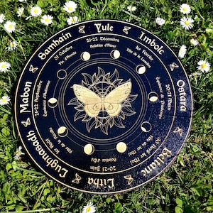 May include: A black wooden wheel with gold accents depicting the eight sabbats of the Wheel of the Year. The wheel features a butterfly design in the center and the names of each sabbat in French and English. The wheel is surrounded by a ring of white daisies.