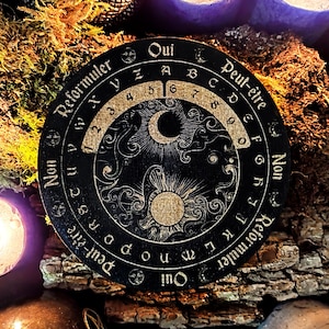 Op de afbeelding: Een zwart houten Ouija-bord met gouden letters en symbolen. Het bord heeft een halve maan en zon ontwerp in het midden. De tekst op het bord luidt "Oui", "Peut-être", "Non" en "Reformuler".