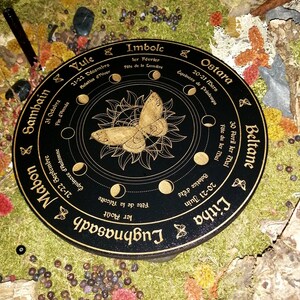 May include: A black circular wooden wheel with gold lettering and a gold butterfly design. The wheel is divided into eight sections, each representing a different pagan holiday. The holidays are: Yule, Imbolc, Ostara, Beltane, Litha, Lughnasadh, Mabon, and Samhain.