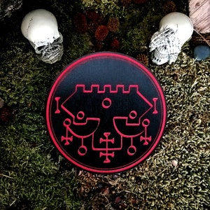 May include: A black and red circular sigil with a complex geometric design. The sigil is surrounded by a red border and is set against a mossy green background.
