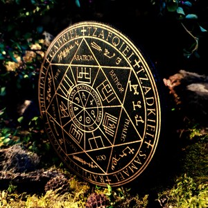 K&ouml;nnte beinhalten: Ein schwarzer runder Untersetzer aus Holz mit einem goldfarbenen Design. Das Design zeigt einen Stern mit einem Kreis in der Mitte und die Worte "SAMAEL RAPHAEL OKIEL+ IZAFIEL" um den Rand herum.