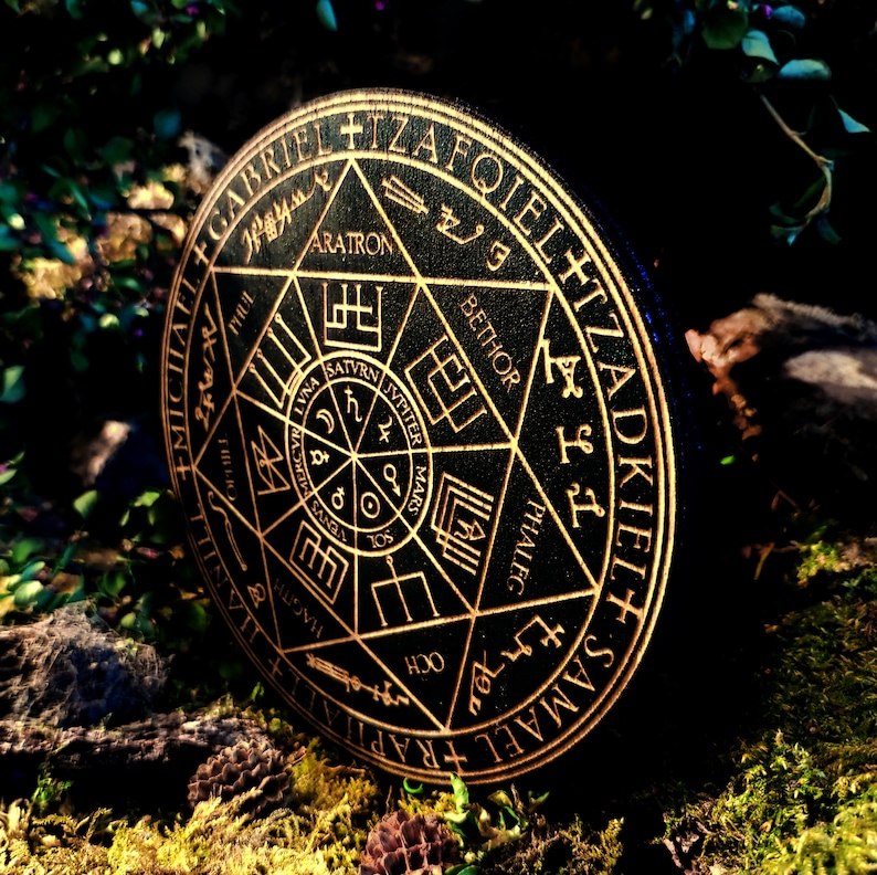 K&ouml;nnte beinhalten: Ein schwarzer runder Untersetzer aus Holz mit einem goldfarbenen Design. Das Design zeigt einen Stern mit einem Kreis in der Mitte und die Worte "SAMAEL RAPHAEL OKIEL+ IZAFIEL" um den Rand herum.