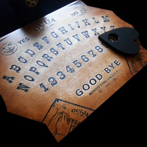 May include: A wooden Ouija board with a black heart-shaped planchette. The board features the letters of the alphabet, numbers 1-9 and 0, and the words "YES", "NO", "GOOD BYE", and "OUIJA SINCE 1891".