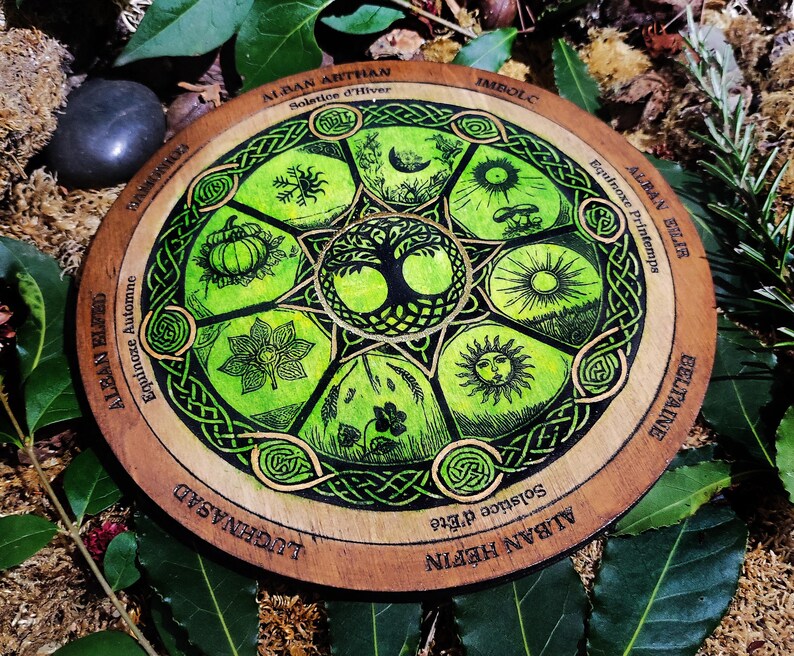 Op de afbeelding: Een houten wiel met een groen en zwart Keltisch knoopontwerp. Het wiel is verdeeld in acht secties, die elk een andere tijd van het jaar in de Keltische kalender vertegenwoordigen. De secties zijn gelabeld met de namen van de festivals: Alban Arthan, Imbolc, Alban Eilir, Beltane, Alban Hefin, Lughnasad, Alban Elfed en Mabon.