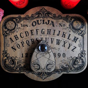 May include: A vintage-style Ouija board made of wood, featuring the alphabet, numbers, and the words "Yes" and "No." The planchette has a clear gem. Ornate black scrollwork frames the board. A bundle of pink sage and two red candles are in the background.