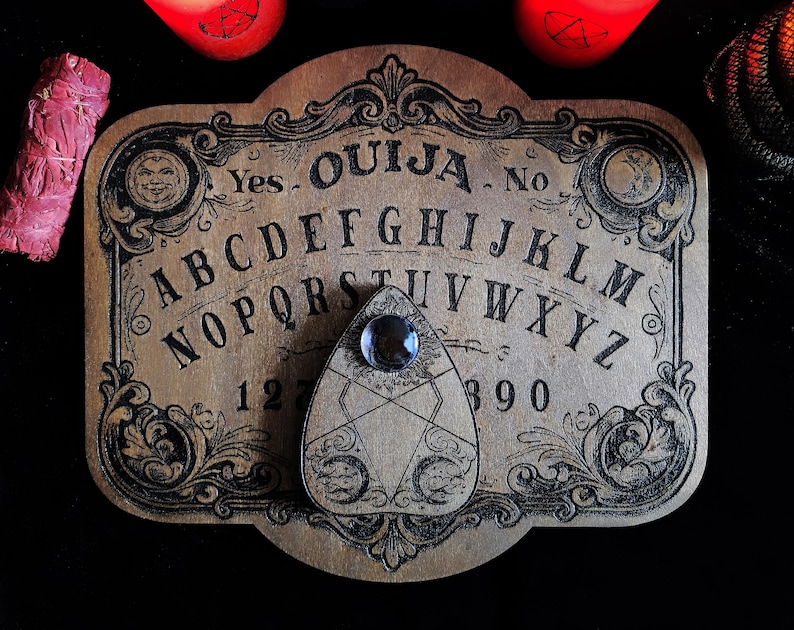 May include: A vintage-style Ouija board made of wood, featuring the alphabet, numbers, and the words "Yes" and "No." The planchette has a clear gem. Ornate black scrollwork frames the board. A bundle of pink sage and two red candles are in the background.