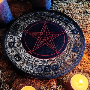 May include: A black wooden circle with a pentagram and goat's head design in red. The circle is surrounded by a ring of symbols and numbers. The circle is on a brown surface with candles.