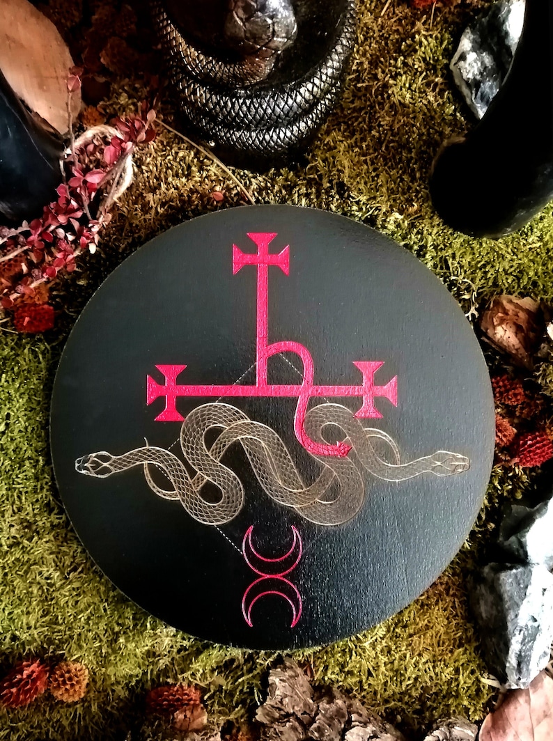 May include: A black circular wooden plaque with a red and gold sigil of a snake wrapped around a cross. The sigil is surrounded by a crescent moon symbol.