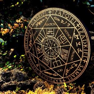 K&ouml;nnte beinhalten: Eine schwarze runde Holzplakette mit goldenen Schriftz&uuml;gen und Symbolen. Die Plakette zeigt einen siebenzackigen Stern mit den Namen von Engeln um den Umfang. Die Mitte des Sterns enth&auml;lt einen kleineren Kreis mit den Namen der Planeten und Symbolen.