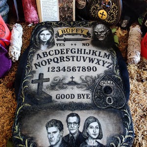 May include: A decorative Ouija board with the word "Buffy" at the top, featuring portraits and symbols. The board includes the alphabet, numbers, and the words "Yes," "No," and "Good Bye."