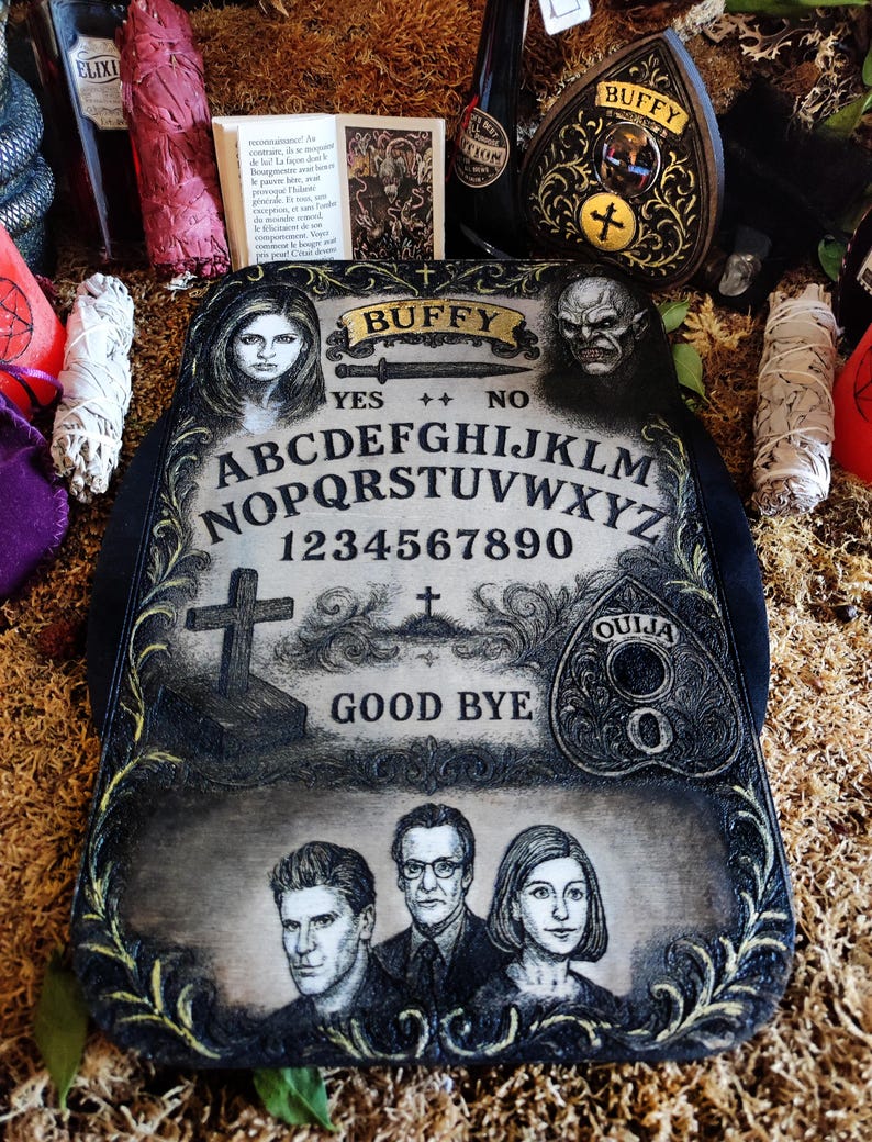 May include: A decorative Ouija board with the word "Buffy" at the top, featuring portraits and symbols. The board includes the alphabet, numbers, and the words "Yes," "No," and "Good Bye."