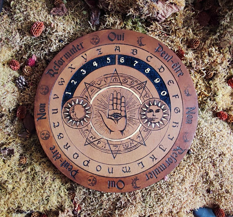 May include: A wooden Ouija board with a black and gold design. The board features a hand with an eye in the center, surrounded by a star and sun symbols. The board has the letters A-Z, numbers 1-9, and the words "Oui", "Non", "Peut-&ecirc;tre", and "Reformuler" written around the edge.