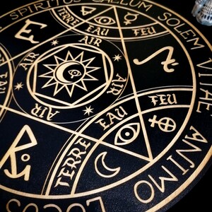 May include: A black wooden decorative round with gold-colored etched symbols and text. The design includes a central star with an eye, crescent moons, and various symbols. The text includes words like "SPIRITUS", "CAELUM", "SOLEM", "TERRE", "EAU", "FEU", and "ANIMO".