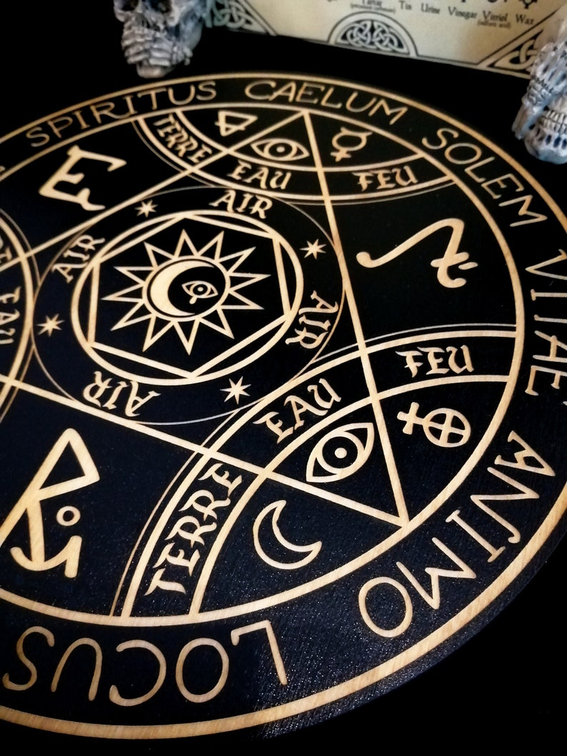 May include: A black wooden decorative round with gold-colored etched symbols and text. The design includes a central star with an eye, crescent moons, and various symbols. The text includes words like "SPIRITUS", "CAELUM", "SOLEM", "TERRE", "EAU", "FEU", and "ANIMO".