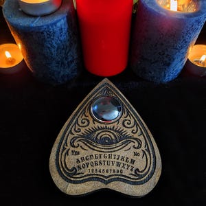 May include: A wooden Ouija board with engraved letters, numbers, and a planchette. The board is surrounded by lit candles of various sizes and colors, including blue, red, and small tea lights, creating a mystical ambiance.