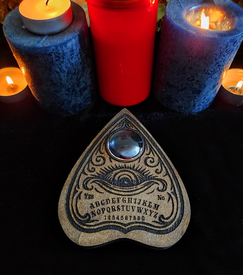 May include: A wooden Ouija board with engraved letters, numbers, and a planchette. The board is surrounded by lit candles of various sizes and colors, including blue, red, and small tea lights, creating a mystical ambiance.