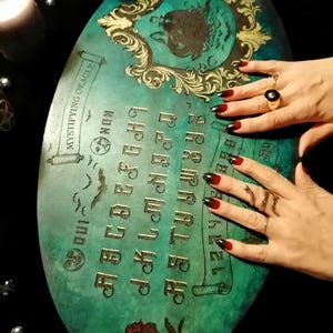 May include: Oval teal Ouija board with gold lettering and a decorative frame. The board features the alphabet, numbers, and the words "Mystifying Oracle". A red rose is at the bottom. Hands with red and black nail polish and gold rings are touching the board.