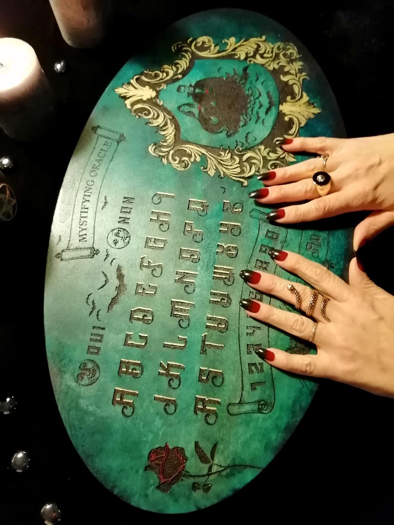 May include: Oval teal Ouija board with gold lettering and a decorative frame. The board features the alphabet, numbers, and the words "Mystifying Oracle". A red rose is at the bottom. Hands with red and black nail polish and gold rings are touching the board.