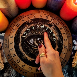 May include: A wooden Ouija board with a black and white eye design and numbers 1 through 9. The board has the words "Oui", "Peut-&ecirc;tre", "Non", "Reformuler", "Oui", "Reformuler", "Non", "Peut-&ecirc;tre", "Oui", "XYZABCDEFGHIJKLMNOPQRSTUVWXYZ" printed around the edge.