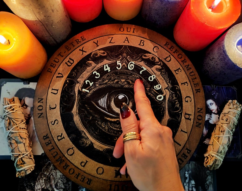 May include: A wooden Ouija board with a black and white eye design and numbers 1 through 9. The board has the words "Oui", "Peut-&ecirc;tre", "Non", "Reformuler", "Oui", "Reformuler", "Non", "Peut-&ecirc;tre", "Oui", "XYZABCDEFGHIJKLMNOPQRSTUVWXYZ" printed around the edge.